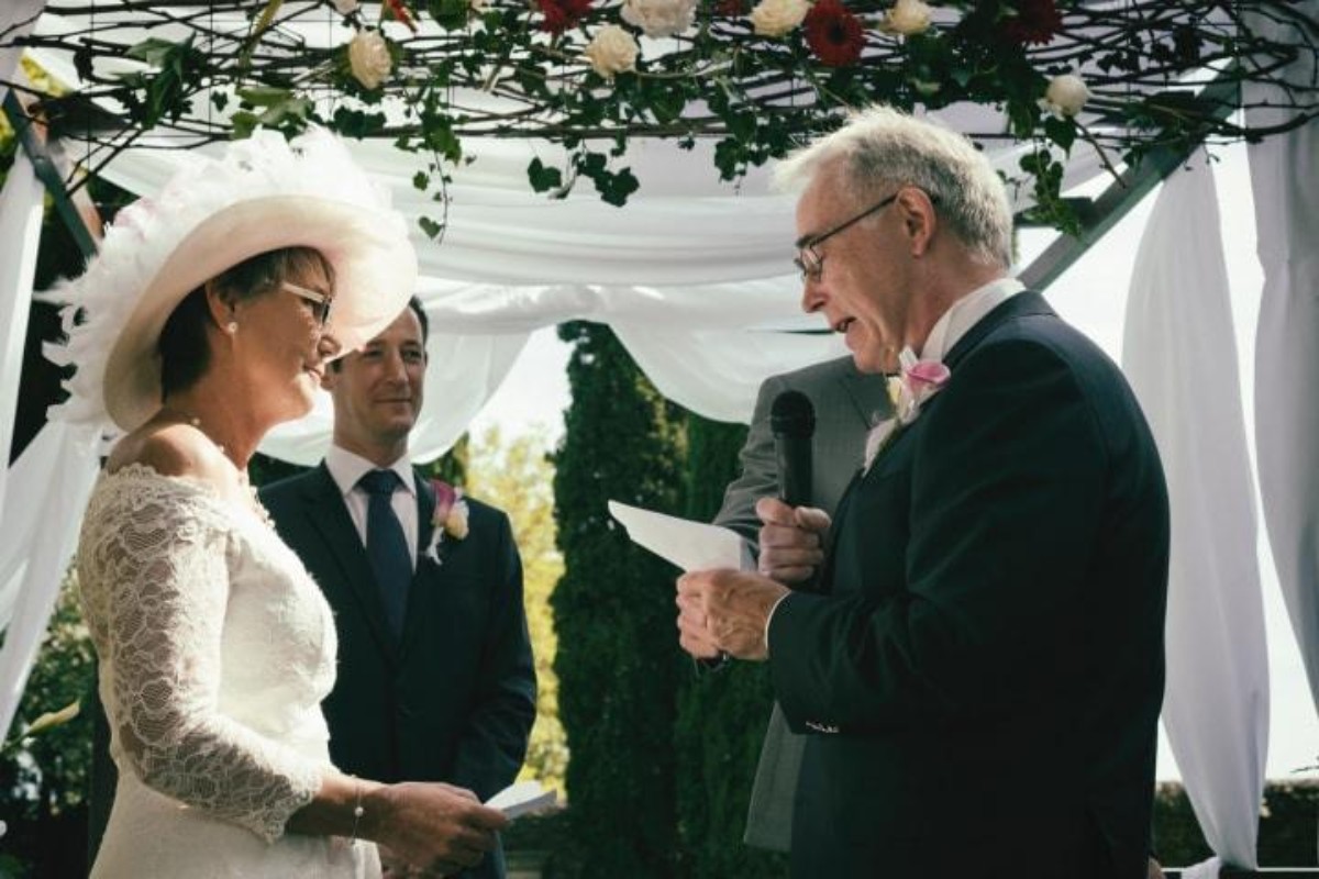 Un couple souriant échange ses vœux sous une arche fleurie lors de leurs noces de porphyre, célébrant 33 ans de mariage entourés de proches.